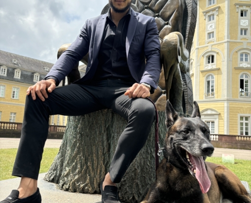 Callboy Alex Léon, scharf gekleidet in einem dunklen Anzug und mit Sonnenbrille, sitzt auf der rauen Oberfläche einer Baumstumpfskulptur. Zu seinen Füßen ruht ein großer, kräftig aussehender dunkler Hund - aufmerksam und ruhig an seiner Seite. Hinter ihnen erhebt sich ein verziertes gelbes Gebäude, dessen Architektur durch hohe Bogenfenster und komplizierte Details gekennzeichnet ist. Der wolkenverhangene Himmel wirft ein diffuses Licht über die Szene und verleiht ihr eine stimmungsvolle Atmosphäre, die im Kontrast zur reichen Fassade des Gebäudes steht.
