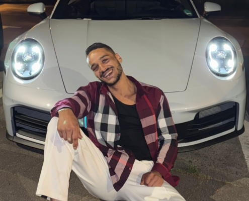 Eine kreative Fotobeschreibung könnte so lauten: Callboy Steve sitzt auf dem Asphalt unter dem Nachthimmel, sein freundliches Lächeln wird vom Scheinwerferlicht eines weißen Sportwagens hinter ihm erhellt. Er trägt ein kariertes Hemd und eine knackige weiße Hose, eine Mischung aus lässigem Selbstbewusstsein und kühnem Stil. Der Parkplatz ist dunkel, bis auf die Lichtflecken, die das Auto wirft, dessen schlanke Silhouette sich von den Schatten abhebt. Steve sieht entspannt und selbstbewusst aus, die Beine lässig ausgestreckt, während er für eine unvergessliche Aufnahme posiert.
