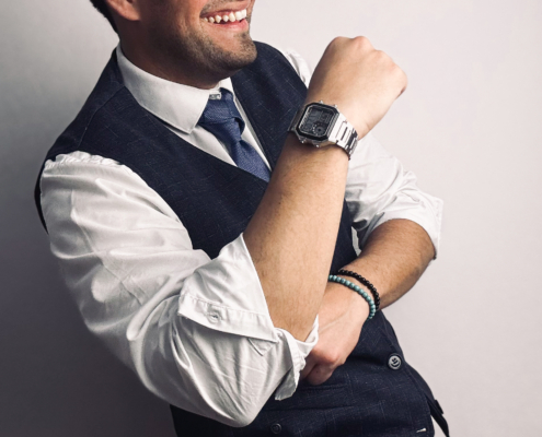Callboy Maxim steht vor einem hellen Hintergrund und strahlt Selbstvertrauen und Charme aus. Er trägt ein frisches weißes Hemd, eine dunkle Weste und eine elegante Krawatte. Eine elegante Uhr ziert sein Handgelenk und verleiht ihm einen Hauch von Eleganz. Maxim lächelt herzlich, beugt den Arm am Ellbogen und streckt energisch die Faust in die Höhe, während er sich leicht zur Seite neigt – seine entspannte Haltung und sein lebhafter Ausdruck strahlen Freundlichkeit und Selbstbewusstsein aus.