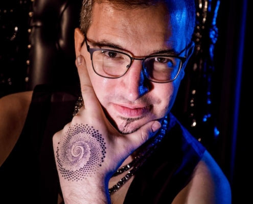 Es klingt, als würden Sie das Porträt eines Mannes mit markanten Merkmalen beschreiben - Brille, Spitzbart und ein detailliertes Spiralpunkt-Tattoo auf dem Handrücken. Das Bild ist kunstvoll mit blauen und rosafarbenen Tönen beleuchtet, was ihm eine moderne oder vielleicht Cyberpunk-Ästhetik verleiht. Möchten Sie mit dieser Beschreibung etwas Bestimmtes tun, z. B. sie in einen Kunstspruch verwandeln oder den Stil eines Künstlers identifizieren? Lassen Sie mich wissen, wie ich Ihnen helfen kann!.