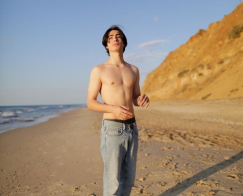 Ein junger Mann mit mittellangem, dunklem Haar steht barfuß auf dem goldenen Sand in der Nähe des Meeres, seine hellblauen Jeans sind bis knapp über die Knöchel gerollt. Hinter ihm ragen zerklüftete Klippen in den strahlenden Himmel, der von sanften Wolken durchzogen ist. Callboy Aslan strahlt einen mühelosen Charme aus, während er in die Ferne blickt - eine Figur von lässiger Zuversicht vor der heiteren, wilden Landschaft.