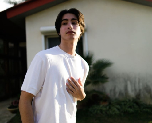 Gewiss! Hier ist eine Beschreibung eines solchen Bildes: Eine junge Person mit mittellangem dunklem Haar steht lässig im Freien und verkörpert eine entspannte Atmosphäre, die an den Stil von Callboy Aslan erinnert. Sie trägt ein einfaches weißes T-Shirt und eine Jeans und strahlt damit mühelose Coolness aus. Hinter ihnen befindet sich ein helles Haus mit einem auffälligen roten Dach, eingebettet in üppiges Grün - Bäume und Büsche umrahmen die Szene und verstärken die entspannte Atmosphäre. Das gedämpfte Sonnenlicht verleiht der gesamten Szenerie Wärme. Wenn Sie möchten, dass ich Ihnen bei der Erstellung dieses Bildes helfe oder Details weiter verfeinere (z. B. Pose oder Gesichtsausdruck), lassen Sie es mich einfach wissen!.