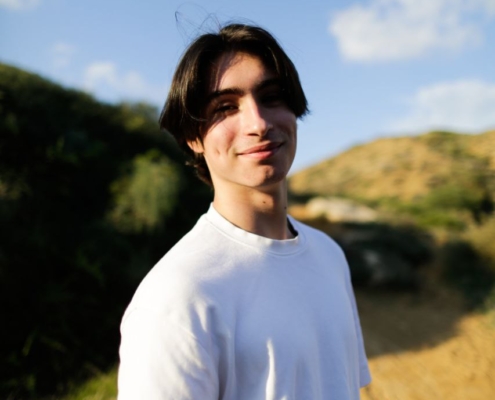 Ein junger Mensch mit dunklem Haar steht selbstbewusst im Freien und trägt ein einfaches weißes T-Shirt. Sie lächelt warm in die Kamera und verkörpert einen unbeschwerten, sorglosen Geist, der an den Callboy Aslan erinnert. Der Hintergrund zeigt üppiges grünes Laub und Bäume neben einem sonnenbeschienenen Feldweg, der sich in die Ferne windet. Der weite, blaue Himmel mit seinen vereinzelten Wolken unterstreicht die entspannte Sommeratmosphäre der Szene.
