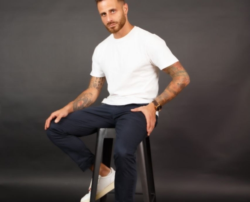 Bruno, der Callboy, sitzt lässig auf einem schwarzen Hocker vor einer schlichten, dunklen Kulisse. Seine Erscheinung ist auffällig - unter den Ärmeln seines knackigen weißen T-Shirts sind tätowierte Arme zu sehen. Er trägt eine taillierte dunkle Hose und makellose weiße Turnschuhe. Brunos Haltung ist entspannt: Sein rechter Arm ruht bequem auf seinem Knie, und er lächelt leicht und selbstbewusst, was auf einen leichten Charme schließen lässt. Das Gesamtbild ist minimalistisch und dennoch überzeugend und lenkt die Aufmerksamkeit auf seine ausdrucksstarken Tätowierungen und sein entspanntes Auftreten inmitten der gedämpften Umgebung.