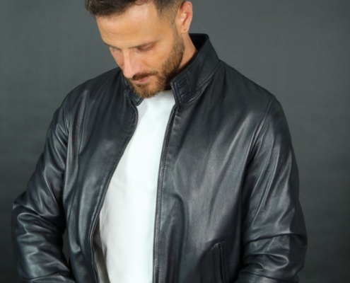 Ein Foto zeigt Callboy Bruno, einen Mann mit kurzen braunen Haaren und Bart, vor einem schlichten dunklen Hintergrund. Er trägt eine schwarze Lederjacke über einem weißen Hemd und blickt nach unten, wobei sein Blick darauf gerichtet ist, die Vorderseite seiner Jacke mit beiden Händen zu richten. Sein Gesichtsausdruck ist nachdenklich oder introspektiv, und die Gesamtstimmung des Bildes ist zurückhaltend und stilvoll.