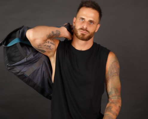 Der tätowierte Callboy Bruno, der ein ärmelloses schwarzes Hemd und eine dunkle Hose trägt, steht mit seiner Jacke über der Schulter vor einem dunklen Hintergrund.