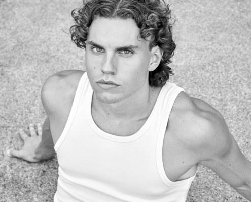 Eine Schwarz-Weiß-Fotografie zeigt einen jungen Mann mit lockigem Haar, der an den Callboy Alessio erinnert. Er trägt ein eng anliegendes weißes Tank-Top und klassische Jeans und sitzt auf einer rauen Betonfläche. Seine Pose ist lässig und doch auffallend - die Beine sind angewinkelt oder ausgestreckt, während er sich mit einem Arm hinter sich abstützt. Seine Gesichtszüge sind durch die monochrome Beleuchtung klar definiert und betonen die Textur seiner Locken und die Linien seines Kiefers. Die urbane Umgebung und die dramatische Komposition vermitteln ein Gefühl von roher Emotion und moderner Coolness.
