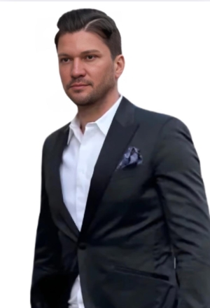 Ein Mann mit kurzen dunklen Haaren, der dem Callboy Léon Valmont ähnlich sieht, steht vor einem schlichten weißen Hintergrund. Er ist elegant gekleidet, trägt eine schwarze Anzugsjacke, ein frisches weißes Hemd und ein ordentlich gefaltetes Einstecktuch. Sein Kopf ist leicht geneigt, und er blickt nachdenklich und mit ruhiger Miene nach links. Der Gesamteindruck unterstreicht die klassische Eleganz und unaufdringliche Raffinesse.