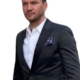 Ein Mann mit kurzen dunklen Haaren, der dem Callboy Léon Valmont ähnlich sieht, steht vor einem schlichten weißen Hintergrund. Er ist elegant gekleidet, trägt eine schwarze Anzugsjacke, ein frisches weißes Hemd und ein ordentlich gefaltetes Einstecktuch. Sein Kopf ist leicht geneigt, und er blickt nachdenklich und mit ruhiger Miene nach links. Der Gesamteindruck unterstreicht die klassische Eleganz und unaufdringliche Raffinesse.
