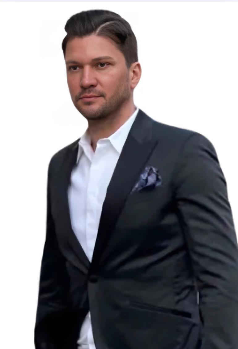 Ein Mann mit kurzen dunklen Haaren, der dem Callboy Léon Valmont ähnlich sieht, steht vor einem schlichten weißen Hintergrund. Er ist elegant gekleidet, trägt eine schwarze Anzugsjacke, ein frisches weißes Hemd und ein ordentlich gefaltetes Einstecktuch. Sein Kopf ist leicht geneigt, und er blickt nachdenklich und mit ruhiger Miene nach links. Der Gesamteindruck unterstreicht die klassische Eleganz und unaufdringliche Raffinesse.
