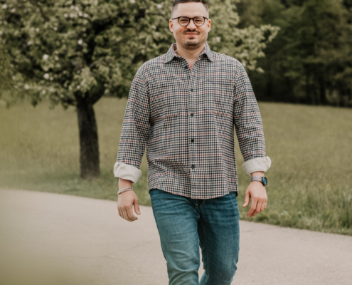 Der Callboy Liam, bekleidet mit einem karierten Hemd und Jeans, geht selbstbewusst über einen gepflasterten Weg im Freien. Er trägt eine Brille, die sein Gesicht nachdenklich umrahmt. Der Weg ist von saftig grünem Gras gesäumt, und belaubte Bäume bilden eine ruhige Kulisse. Das Sonnenlicht fällt durch das Laub und wirft sanfte Schatten, während er vorwärts schlendert und sich in der natürlichen Umgebung entspannt und wohl zu fühlen scheint.