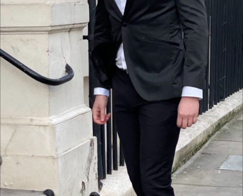 Sie beschreiben eine modische Stadtszene: Ein Mann, elegant gekleidet in schwarzer Anzugsjacke und -hose mit weißem Hemd und schwarzen Halbschuhen, geht einen städtischen Bürgersteig entlang. Die Kulisse ist ein elegantes weißes Gebäude mit einem schmiedeeisernen Geländer. Der Kontrast zwischen seiner klaren, formellen Kleidung und den klassischen architektonischen Details deutet auf Raffinesse hin - vielleicht in einem eleganten Viertel oder Geschäftsviertel. Wenn Sie eine weitere Beschreibung oder Analyse benötigen oder dieses Bild für kreative Texte oder andere Zwecke visualisieren möchten, lassen Sie es mich wissen!.