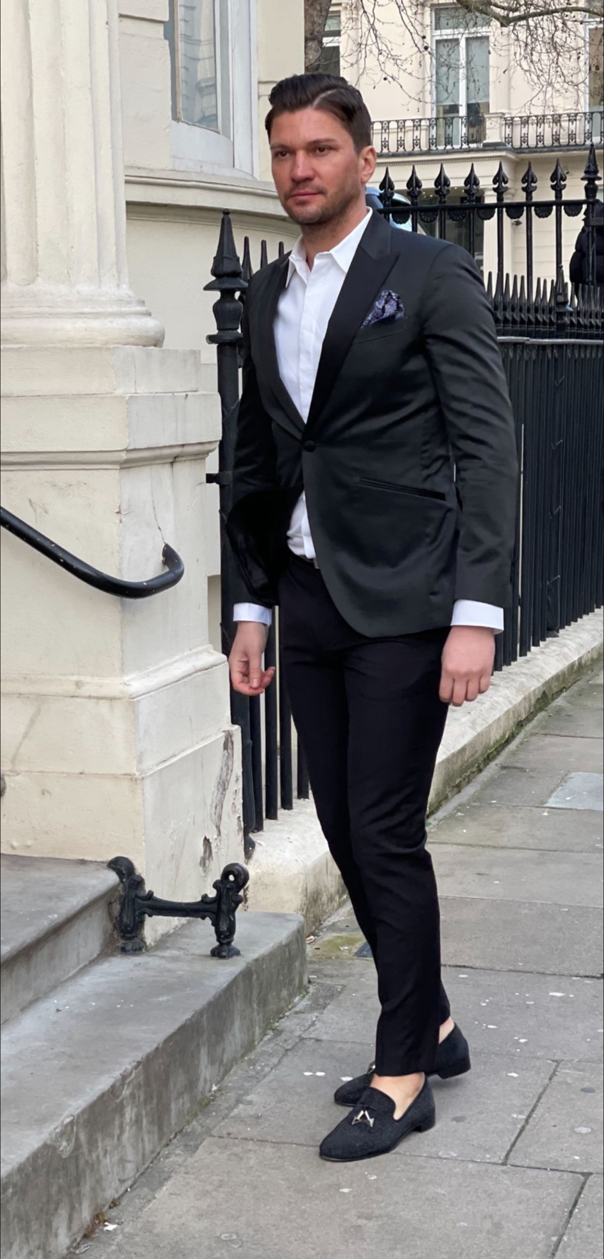 Sie beschreiben eine modische Stadtszene: Ein Mann, elegant gekleidet in schwarzer Anzugsjacke und -hose mit weißem Hemd und schwarzen Halbschuhen, geht einen städtischen Bürgersteig entlang. Die Kulisse ist ein elegantes weißes Gebäude mit einem schmiedeeisernen Geländer. Der Kontrast zwischen seiner klaren, formellen Kleidung und den klassischen architektonischen Details deutet auf Raffinesse hin - vielleicht in einem eleganten Viertel oder Geschäftsviertel. Wenn Sie eine weitere Beschreibung oder Analyse benötigen oder dieses Bild für kreative Texte oder andere Zwecke visualisieren möchten, lassen Sie es mich wissen!.