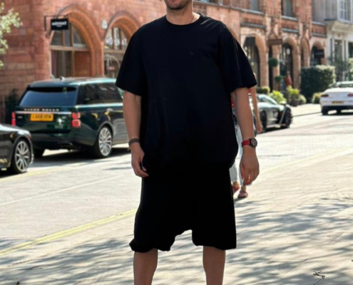 Es klingt so, als würden Sie ein Bild beschreiben, auf dem ein leger gekleideter Mann in einer sonnigen Stadtstraße steht. Er trägt eine Sonnenbrille, ein schwarzes T-Shirt und schwarze Shorts. Hinter ihm befinden sich rote Backsteingebäude und mehrere geparkte Autos. Die Gesamtatmosphäre deutet auf einen hellen Tag hin, vielleicht in einer Stadt oder einem Wohnviertel. Wenn Sie möchten, dass ich eine künstlerische Beschreibung erstelle, Ihnen Outfitvorschläge für ähnliches Wetter mache oder Ihnen bei etwas Speziellem in Bezug auf diese Szene helfe (z. B. beim Schreiben von Alt-Text oder beim Verfassen einer Geschichte auf der Grundlage des Bildes), lassen Sie es mich bitte wissen!.