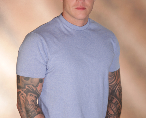 Ein Mann mit kurzen Haaren und sichtbaren Armtattoos steht selbstbewusst da und hat die Hände in den Taschen seiner hellblauen Jeans. Er trägt ein weich geschnittenes hellblaues T-Shirt, das sein lässiges und doch stilvolles Aussehen unterstreicht. Der Hintergrund besteht aus einem sanften Farbverlauf in Braun- und Beigetönen, der der Szene eine elegante Wärme verleiht, die an professionelle Studioporträts erinnert. Seine entspannte Haltung und seine charismatische Präsenz vermitteln die schicke, modische Ausstrahlung, die oft mit Callboy Andreas in Verbindung gebracht wird - inspiriert von modernem Lifestyle und Flair.