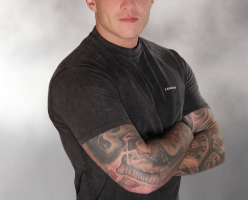 Andreas steht selbstbewusst vor einem glatten grauen Studiohintergrund. Sein kurzgeschnittenes Haar betont seine kantigen Gesichtszüge, und verschlungene Tattoos schlängeln sich entlang seiner beiden Arme - sichtbar, wenn er sie vor der Brust verschränkt. Das schwarze T-Shirt liegt eng an und unterstreicht seinen gut gebauten Körper, während die hellen Jeans einen Kontrast und einen lässigen Touch bieten. Sein Gesichtsausdruck ist ernst und doch sympathisch - ein professioneller Callboy, der sich vor der Kamera wohlfühlt und sowohl Stärke als auch Sympathie ausstrahlt.