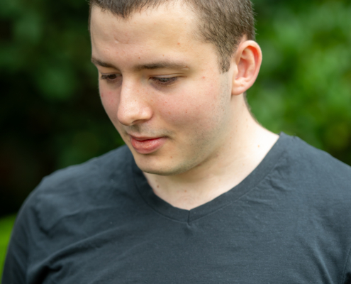 Ein junger Mann mit kurzen braunen Haaren, bekleidet mit einem schwarzen T-Shirt mit V-Ausschnitt, steht da und blickt nachdenklich nach unten. Der Hintergrund ist eine kunstvolle Unschärfe aus Grün - weich fokussierte Blätter und Laub -, die die Szene mit einem Gefühl von Geheimnis und Ruhe umhüllt. Diese heitere Szenerie umrahmt die nachdenkliche Stimmung des Callboys Alessio und unterstreicht sowohl seinen introspektiven Ausdruck als auch die Faszination der unbekannten Umgebung um ihn herum.