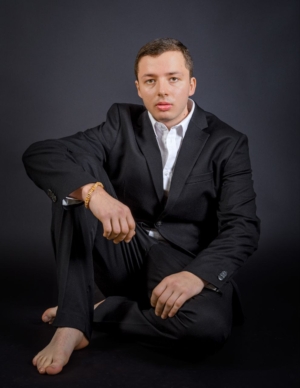 Callboy Tim, in schwarzem Anzug und weißem Hemd, sitzt barfuß auf dem Boden, hat ein Knie hochgezogen und starrt ernst in die Kamera.