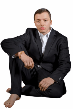 Callboy Tim, barfuß, in schwarzem Anzug und weißem Hemd, sitzt auf dem Boden, ein Knie aufgeschlagen, den Arm darauf gestützt und in die Kamera blickend.