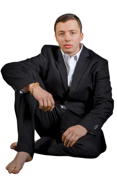 Callboy Tim, barfuß, in schwarzem Anzug und weißem Hemd, sitzt auf dem Boden, ein Knie aufgeschlagen, den Arm darauf gestützt und in die Kamera blickend.