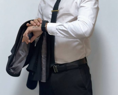 Ein Mann mit weißem Hemd und schwarzer Krawatte, wie Callboy Massimo, hält eine Anzugsjacke und kontrolliert seine Uhr vor einem hellen Hintergrund.