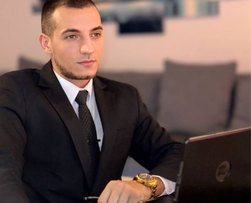 Ein Mann im Anzug wie Callboy Massimo sitzt an einem Tisch mit einem Laptop, in der Hand einen Kaffee, konzentriert in einem modernen Wohnzimmer mit Kunst.