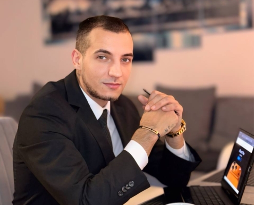 Ein Mann im Anzug, wie Callboy Massimo, sitzt mit Kaffee und Tablet an einem Tisch, die Hände gefaltet, in einem modernen Wohnzimmer mit Kunst.