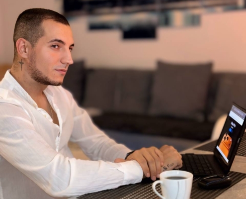 Callboy Massimo, kurzhaarig und im weißen Hemd, sitzt mit Laptop und Kaffee an einem Tisch und blickt zur Seite; hinter ihm die Couch.