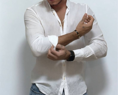 Callboy Massimo steht in einem weißen Button-Down-Hemd und einer blauen Jeans vor einem schlichten weißen Hintergrund und schaut nach unten, während er seine Ärmel hochkrempelt.