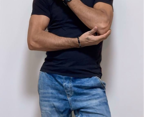 Ein Mann in einem schwarzen T-Shirt, zerrissenen Bluejeans und einer Smartwatch steht an einer weißen Wand, die Hand am Kinn, den anderen Arm verschränkt.