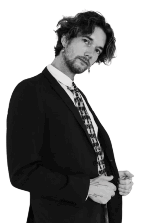 Ein Mann mit gewelltem Haar, der die Eleganz des Callboy Eros ausstrahlt, rückt seinen Anzug und seine gemusterte Krawatte vor einem strahlend weißen Hintergrund zurecht.