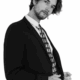 Ein Mann mit gewelltem Haar, der die Eleganz des Callboy Eros ausstrahlt, rückt seinen Anzug und seine gemusterte Krawatte vor einem strahlend weißen Hintergrund zurecht.