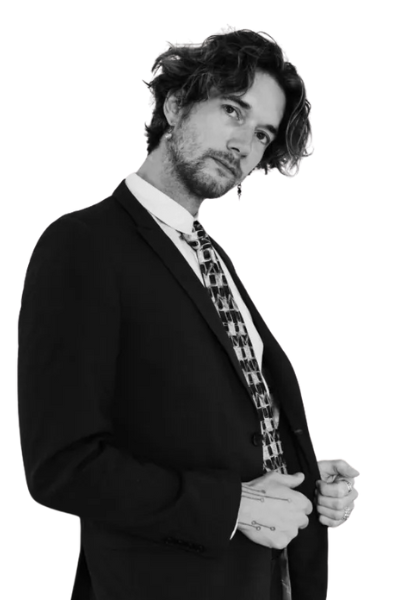 Ein Mann mit gewelltem Haar, der die Eleganz des Callboy Eros ausstrahlt, rückt seinen Anzug und seine gemusterte Krawatte vor einem strahlend weißen Hintergrund zurecht.
