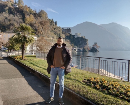 Ein Mann in dunkler Jacke und Jeans steht an einem sonnigen Tag an einem See, hinter ihm Berge und Bäume, in der Nähe Blumen und eine Palme.
