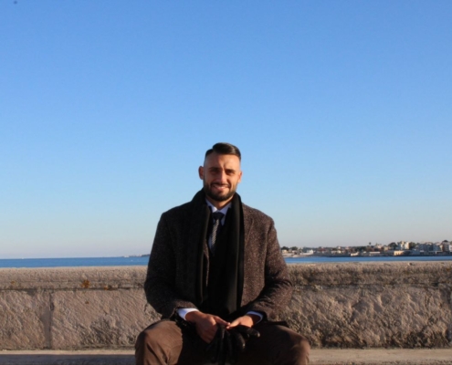 Ein Mann in Mantel und Schal sitzt auf einem Steinsockel am Meer und grinst in die Kamera. Der blaue Himmel und entfernte Gebäude vervollständigen die Szene.