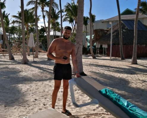 Mann in Badehose steht barfuß an einem Sandstrand mit Palmen und hält ein Telefon in der Hand; Sonnenliegen, Handtücher und tropische Gebäude.