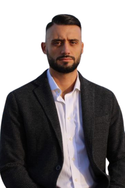 Callboy Jacques mit kurzen dunklen Haaren und einem Bart posiert selbstbewusst in einem weißen Hemd und einem karierten Blazer auf weißem Hintergrund.