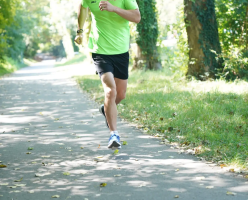 Ein Mann in einem hellgrünen Hemd und schwarzen Shorts joggt über einen sonnenbeschienenen, von Bäumen gesäumten und von Grünflächen umgebenen Parkweg.