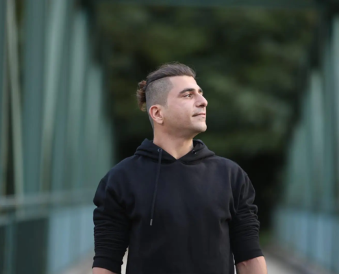 Eine Person mit kurzen dunklen Haaren und schwarzem Kapuzenpulli steht auf einer Brücke und schaut nachdenklich nach oben; dahinter verschwommene Bäume und Strukturen.