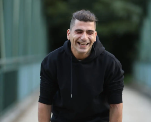 Ein Mann in schwarzem Kapuzenpulli und blauer Jeans steht lächelnd auf einer Brücke und lehnt sich nach vorne, während verschwommenes Grün den Callboy Rico umgibt.