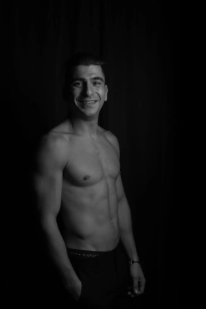 Ein Mann ohne Hemd, der dem Callboy Rico ähnelt, lächelt in die Kamera. Vor einem dunklen Hintergrund werden sein durchtrainierter Körper und seine Gesichtszüge in Schwarz-Weiß hervorgehoben.