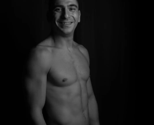 Ein Mann ohne Hemd, der dem Callboy Rico ähnelt, lächelt in die Kamera. Vor einem dunklen Hintergrund werden sein durchtrainierter Körper und seine Gesichtszüge in Schwarz-Weiß hervorgehoben.