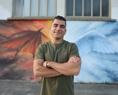 Ein Mann mit kurzen dunklen Haaren und grünem T-Shirt steht mit verschränkten Armen lächelnd vor einem Wandbild-Rico zwischen einem roten Drachen und einem weißen Engelsflügel.