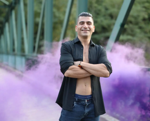 Callboy Rico steht mit verschränkten Armen und einem Lächeln auf einer Brücke, trägt ein offenes schwarzes Hemd und Jeans, während hinter ihm lila Rauch aufsteigt.