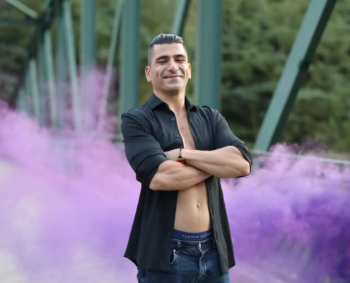 Callboy Rico, dunkles Haar, offenes schwarzes Hemd und Jeans, steht auf einer Brücke inmitten von lila Rauchschwaden und Bäumen.