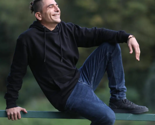 Ein Mann in schwarzem Kapuzenpulli und Jeans lehnt sich auf einem Geländer im Freien und lächelt nach oben. Rico hat ein Bein hochgelegt, der Arm ruht, dahinter ein grüner Fleck.