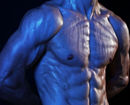 Ein hemdsärmeliger, muskulöser Mann mit glänzender Haut posiert unter blauem Licht vor einem dunklen Hintergrund; der Kopf von Callboy Samuel ist teilweise abgeschnitten.