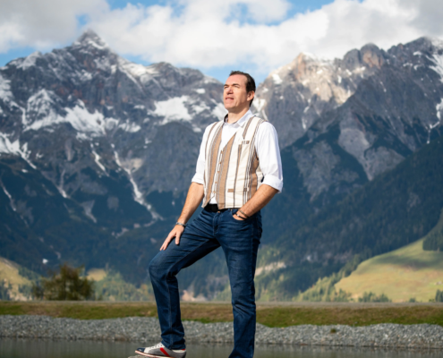 Ein Mann in Jeans, weißem Hemd und gestreifter Weste steht auf einem Felsen an einem spiegelnden See, mit schneebedeckten Gipfeln und Wolken im Hintergrund.