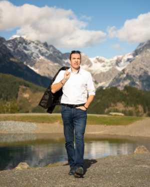 Selbstbewusster Mann in weißem Hemd und Jeans, Jacke über die Schulter geworfen; Berge, See und Bäume im Hintergrund unter teilweise bewölktem Himmel.