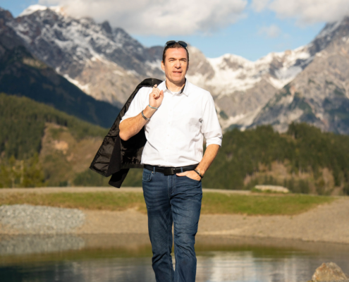 Selbstbewusster Mann in weißem Hemd und Jeans, Jacke über die Schulter geworfen; Berge, See und Bäume im Hintergrund unter teilweise bewölktem Himmel.